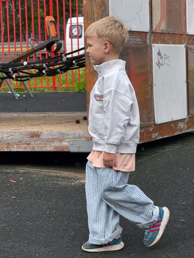 A young boy with blonde hair, wearing a light grey zippered sweatshirt over a peach t-shirt and light blue and white striped pants, walks to the right. He is wearing blue and red sneakers. The boy is on a playground with dark grey asphalt covering the ground. In the background, there is a wooden playground structure with some metal elements and what appears to be a red fence. Pinecones are visible on the wooden surface of the playground. The location is Bylovo, Russia.