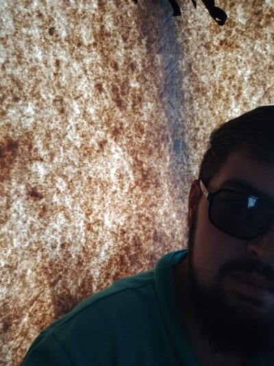 The image shows a close-up, low-angle view of a person wearing sunglasses and a teal-colored collared shirt. The person has dark hair and a beard. They are positioned on the right side of the frame, with their face partially obscured by shadow and the sunglasses. The background is a textured surface with a pattern of brown and white speckles, appearing to be a fabric or canvas material, possibly a tent or similar structure. There are some dark, abstract shapes visible at the top edge of the image, which could be part of the structure or the environment. The lighting suggests an indoor or shaded outdoor setting. The image was captured in Dagestanskiye Ogni, Russia.