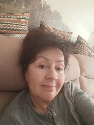 An adult woman with dark, slightly disheveled hair is the central subject of this close-up selfie. She has a warm skin tone and a gentle, closed-mouth smile, looking directly at the camera. She is seated on a plush, light brown upholstered sofa, which features deep cushions and visible seams.
Behind her, several throw pillows display a distinct geometric pattern in shades of orange, brown, grey, and white, featuring zigzags and stripes. The wall behind the sofa is adorned with a large mural or wallpaper depicting a muted, hazy landscape of trees. On the right side of the mural, a faint outline of a tall structure resembling the Eiffel Tower is visible against a lighter, misty sky, suggesting a Parisian theme.
The woman is wearing a grey, ribbed long-sleeved top with a henley-style neckline, fastened by three visible snap buttons on her left side. The activity is a relaxed selfie capture in an indoor setting, likely a living room. The lighting appears to be soft and ambient, suggesting daytime. Her expression conveys a sense of calm and contentment. There is no visible text in the image.