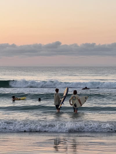 In Wrightsville Beach, United States of America, at dusk, two young males stand in the shallow ocean with surfboards. The male on the left faces away from the camera, holding a surfboard with blue, orange, and white accents. He wears a light grey hoodie and dark pants. The male on the right, also facing away, holds a white surfboard with black markings. He is dressed in a light-colored t-shirt with a graphic design and denim shorts. In the middle distance, a person on a yellow flotation device rests in the water. A second person is partially submerged, with only their head visible, to the left of the two surfboarders. A wave breaks in the background, and other surfers are visible in the distance. The sky displays a soft gradient of peach and pink, with a line of gray clouds near the horizon. The water reflects the colors of the sky. The overall scene captures a serene evening at the beach.