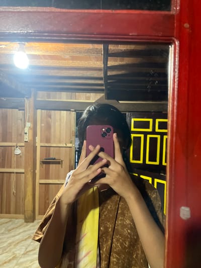 The media file is a slightly distorted, vertically oriented selfie taken in a room with wooden walls. A person, whose face is mostly obscured by a phone, is holding a dark pink smartphone to take the picture. The person is wearing a brown garment with a mottled pattern and a yellow tie-dye pattern. In the reflection of the mirror, a light bulb is visible on the left side of the frame, illuminating the room. The background of the reflection shows wooden planks and what appears to be a decorative element with dark brown and yellow rectangular shapes. There is no visible text in the media file. The overall impression is of an informal selfie taken indoors, likely at night due to the artificial lighting. The wooden interior suggests a rustic or perhaps a traditional setting, consistent with the provided location context of Turumame, Indonesia.