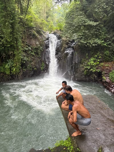 Three men stand on a gray platform overlooking a powerful waterfall. Lush green trees and vegetation surround the rocky terrain. The waterfall cascades down, churning into a vibrant, turbulent pool below. The men are closely positioned, gazing down at the rushing water. Their proximity to the edge suggests they might be contemplating a jump. The scene captures a mix of natural beauty, adventure, and potential danger.