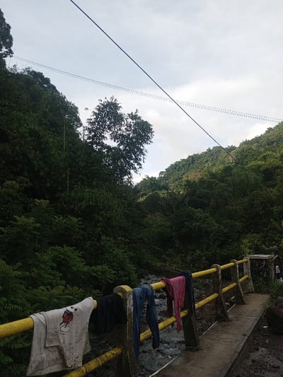 A lush, green valley setting with a small, rushing stream visible below a yellow, metal railing. Clothes hang over the railing to dry. The hillside is thick with vegetation, rising on either side. Power lines stretch across the sky. The day is overcast, the sky a pale grey. A concrete bridge is visible.