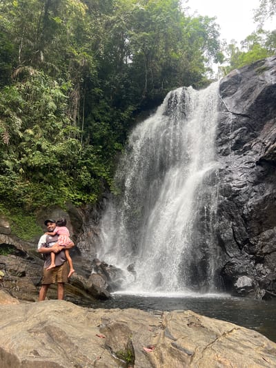 In Lankagama, Sri Lanka, a man stands in front of a cascading waterfall, holding a child. The man, wearing a dark t-shirt and shorts, is looking towards the camera with a smile. The child, dressed in a pink and white striped dress, is being held by the man, facing away from the camera. The waterfall, a prominent feature of the scene, plunges down a rocky cliff face, surrounded by lush green foliage. The area appears to be a natural, tropical environment, with rocks and water forming the foreground and the waterfall and jungle forming the background. The lighting suggests an overcast day.