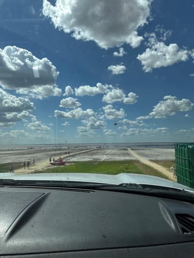 The image is a view from inside a vehicle looking out at a salt lake in Bursol, Russia. The sky is bright blue with fluffy white clouds. In the distance, a vast expanse of salt flats stretches out, dotted with numerous people. A dirt path leads towards the lake, and a red and white car is parked on the side of the path. Green vegetation lines the edge of the salt flats closest to the vehicle. A large green cylindrical object, possibly a dumpster, is visible on the right side of the image. The foreground shows the dashboard of the vehicle.
