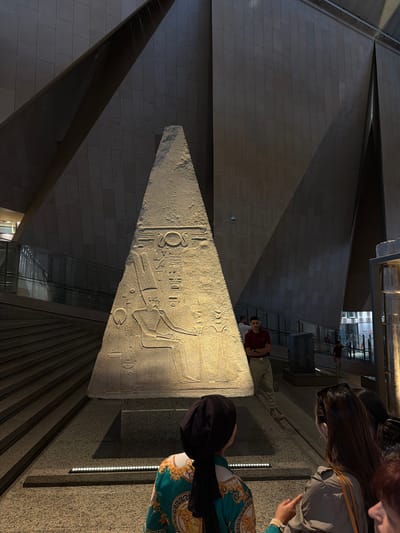 The image shows a group of people in a museum or gallery, likely in Kafrat Nassar, Egypt. The main subject is a large, triangular stone artifact with intricate carvings, resembling an obelisk or stele, illuminated by directional lighting. The artifact features hieroglyphic inscriptions and a relief of a figure, possibly a pharaoh or deity.

Several people are visible, some standing and observing the artifact, while others are partially visible at the edges of the frame. One person in the foreground is wearing a green garment with a gold pattern and a black headscarf. Another person in the background is wearing a maroon shirt and khaki pants. The setting appears to be an indoor space with modern architectural elements, including geometric walls, staircases, and glass partitions, suggesting a museum or cultural exhibition.

The lighting creates dramatic shadows, highlighting the textures of the stone and the carvings. The time of day is indeterminate due to the indoor setting, but the lighting suggests it could be evening or that artificial illumination is solely used. No specific weather conditions are discernible. The overall mood is one of historical appreciation and quiet observation. There is no visible text in the image.