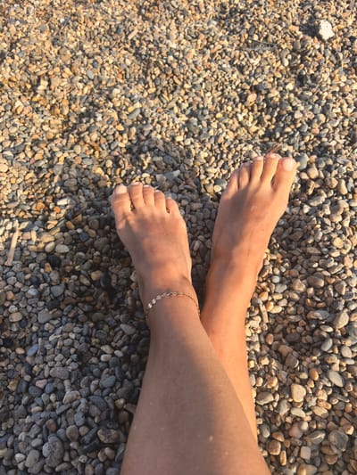 A pair of bare feet rest on a bed of multi-colored pebbles, illuminated by warm sunlight that casts faint shadows. The feet are positioned with the soles facing upwards and crossed at the ankles. A delicate gold anklet, adorned with small circular charms, encircles the left ankle. The surrounding environment consists entirely of densely packed, smooth pebbles in various shades of grey, beige, and brown, with a few scattered pieces of wood. The scene appears to be an outdoor setting, possibly a beach or a rocky shoreline. While the image itself does not provide direct evidence of Myskhako, Russia, the context provided suggests this location.