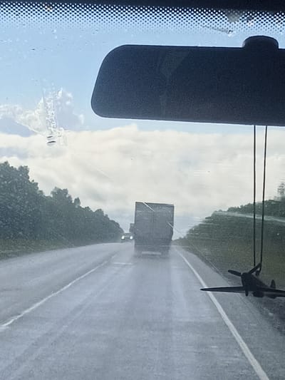 The image depicts a view from inside a vehicle on a wet road, likely during a light rain shower, as evidenced by water streaks on the windshield and the sheen on the asphalt. The scene is set on a highway with dense green foliage lining the sides of the road. In the foreground, hanging from the rearview mirror, is a dark-colored model airplane, possibly a Spitfire. The road ahead is visible, with two trucks in the distance. The truck on the right is larger and appears to be in the foreground relative to the other truck. The truck on the left is smaller and further away. The sky is cloudy with patches of blue visible. The overall impression is of a journey through a rural or semi-rural landscape, with the weather creating a slightly muted and reflective atmosphere. There is no specific indication of the city of Anan'ino, Russia, in the image itself.