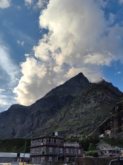 In Damphug, India, a scene of dramatic natural beauty unfolds. Majestic, dark green mountains with rugged, rocky textures dominate the background, their peaks piercing a vast, blue sky filled with voluminous white clouds. The clouds, illuminated by sunlight, cast shadows and add depth to the sky. In the mid-ground, a multi-story building with a gray facade and numerous white-framed windows stands out against the mountain slope. The building appears to be a hotel or guesthouse, with water tanks visible on its roof. Below this building, other smaller structures and trees are visible, suggesting a small settlement nestled within the mountain valley. The overall impression is one of a remote and picturesque location, likely a tourist destination known for its natural scenery.