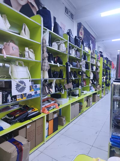 The image shows a retail store interior, specifically a display of handbags and accessories, likely in the city of Ushtobe, Kazakhstan. The store features bright green shelving units that are filled with various styles of handbags, ranging in color from pink and cream to black and brown. Some shelves also contain boxes, plastic bags with items, and promotional signs, one of which says "Скидка" (Discount) in Russian. In the background, there are posters on the wall, one featuring the "VOM The Color Trend Company" logo and another with an image of a woman's face. The floor is tiled with a light-colored, marble-like pattern. There are no people visible in the image. The lighting appears to be artificial, likely from overhead fluorescent lights. The scene is organized and stocked, indicating a functioning retail environment.
