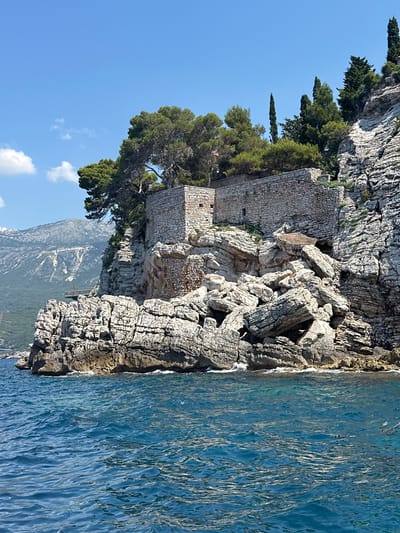 The image shows a fortified structure built on a rocky cliff overlooking the sea. The structure is made of stone and appears to be old. It is surrounded by lush green trees, and the sea is a vibrant blue. The background shows more rocky cliffs and mountains. The sky is clear and blue with a few scattered clouds. The location is the island of Sveti Stefan, near the village of Donji Katun, Montenegro.