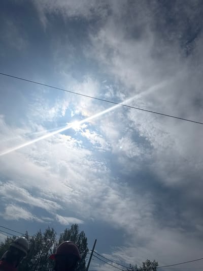 The image depicts a bright, partly cloudy sky with the sun prominently visible, creating a strong lens flare that streaks across the frame. The sun appears high in the sky, suggesting it is midday. Wispy white clouds are scattered across a blue sky, with some darker, more dense clouds present, indicating variable weather. Two black wires, likely power or telephone lines, run diagonally across the sky. In the lower right portion of the image, the tops of trees are visible, their leaves appearing green and full, suggesting a warm season like summer. Two individuals are partially visible at the very bottom, wearing hard hats. One hard hat is red and the other is white and gray. The perspective is looking upwards from ground level. The scene appears to be outdoors, possibly in a residential or semi-rural area, given the presence of trees and utility wires. No text is visible in the image.