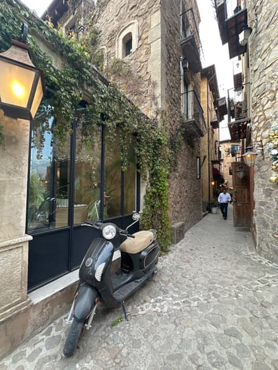 A tilted street scene showcases stone buildings adorned with ivy. A black scooter rests on the cobblestone path near a building with a large glass window. Further down the lane, a person in a white shirt walks away. Streetlights hang above, and decorations are visible near the top of the frame, suggesting a festive atmosphere. The angle provides a unique perspective of the architecture and urban environment.