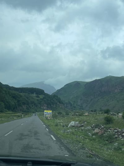 An overcast, cloudy day is captured in this outdoor, eye-level, full-shot photograph, looking down a winding asphalt road through a mountain valley. The road is lined with white dashed lines on the right and a solid white line on the left, and appears to be in fair condition with some darker patches of wear. A few vehicles are visible on the road in the distance, heading away from the viewer. To the right of the road, there is a large billboard with yellow and white signage. The landscape on either side of the road is rugged and mountainous, with steep, green, rocky slopes rising sharply. Dense green trees cover the lower portions of the mountains. The valley floor is covered with rocks and sparse green vegetation. The sky is filled with heavy, gray clouds, suggesting impending rain or a generally damp day. In the background, a prominent mountain peak is visible, partially obscured by clouds. The scene appears to be in Gergeti, Georgia, known for its dramatic mountain landscapes.