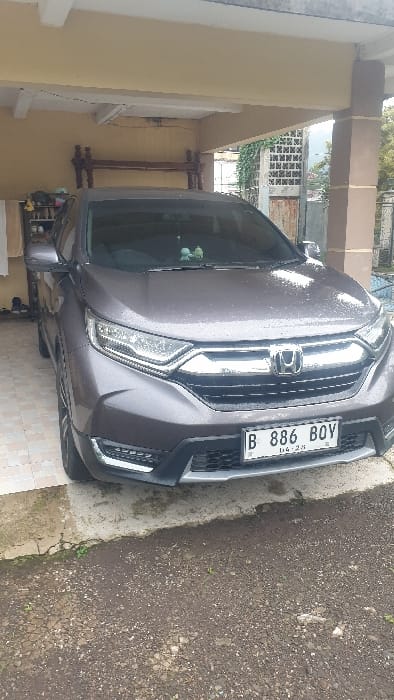 A dark gray Honda CR-V SUV, bearing Indonesian license plate B 886 BOY with an indicated expiration of 04-28, is stationary in a partially covered carport in Gadog, Indonesia. The vehicle's exterior, notably the hood and windshield, exhibits water droplets. The carport features light yellow-painted walls and a white ceiling, supported by brown pillars. The ground beneath the vehicle transitions from a tiled surface on the left to exposed dirt and gravel in the foreground. Along the carport's rear and left walls, a wooden headboard-like object, a small shelf holding shoes, and a white towel hanging on a rack are visible. Beyond the carport's right side, a metal fence with dense green foliage is present, leading to a view of distant hills or mountains under an overcast sky. No individuals, active movements, or interactions are depicted in the scene.