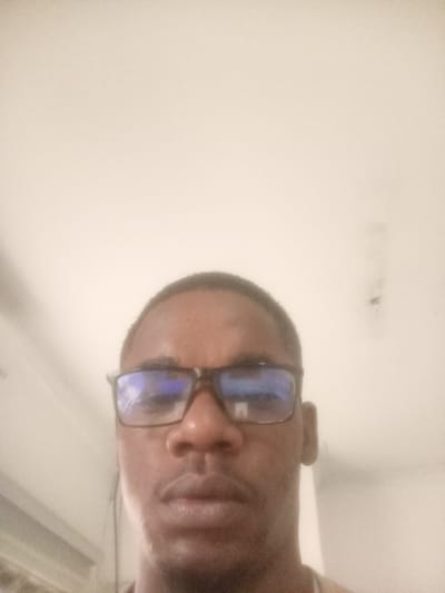 The image is a close-up, selfie-style photograph of a Black man wearing glasses. He is positioned in the lower center of the frame, with his face taking up a significant portion of the image. The background is a plain, light-colored wall, possibly an interior wall. The lighting appears to be overhead, creating some reflections on the lenses of his glasses. The man's expression is neutral. The overall image quality is somewhat grainy, suggesting it was taken with a mobile phone or a low-resolution camera. There is no discernible information to indicate the location in Sudbury, United States of America.