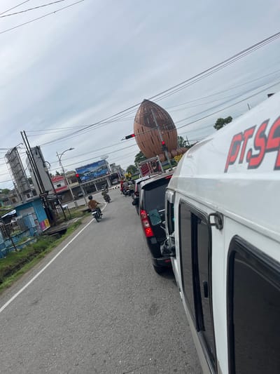 This media file captures a street scene in Sigli, Indonesia, likely during the day given the overall brightness. The perspective is from within a vehicle, looking out towards the road and surrounding environment. The sky appears overcast, suggesting a cloudy or possibly rainy day.
Dominating the foreground on the right is the side of a white vehicle, possibly a van or truck, with black tinted windows. Part of a logo or text is visible on the vehicle's white surface, appearing to be in red, bold lettering. Further down the road, a traffic jam is evident, with several cars and motorcycles stopped. One motorcycle rider, wearing a brown shirt, is clearly visible.
To the left of the road, there's a distinct, large, dome-shaped wooden structure, possibly part of a building or a decorative installation. A red traffic light is illuminated on this structure. Beyond this, there are several buildings of varying sizes and architectural styles, some appearing to be shops or businesses. Prominent billboards are also visible, including one with blue and white imagery of a car, and another with more abstract graphics.
The scene depicts a typical urban street environment with ongoing traffic and commercial activity. The presence of multiple vehicles and pedestrians suggests a bustling area. The overall atmosphere appears to be one of everyday life in a Southeast Asian city, with no overt signs of distress or celebration. The street is paved, and a white line marks the edge of the road. Vegetation, including grass, is visible along the roadside.