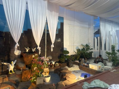 The image depicts an outdoor scene, possibly a garden or event space, with elements suggesting ongoing setup or maintenance. Dominating the foreground and midground is a decorative rock formation with artificial water features and a pond. White fabric drapes hang from the ceiling, creating a tent-like atmosphere.

Several decorative white bird statues are placed on the rocks, with one larger white bird statue with yellow accents positioned on the left. Colorful flowering plants in pots add vibrancy to the scene. The backdrop features a painted mural of a sky with clouds and rocky structures.

In the midground, two individuals are present. One person is seated on the ground, appearing to be working with materials. Another individual is also seated, looking towards the ground. Various objects are scattered around, including a white plastic tub, ropes, a blue plastic tray, and tools, indicating some form of assembly or preparation. The flooring in the foreground appears to be a reddish-brown material, possibly brick or tile. The overall lighting suggests it is either late afternoon or evening. The image is identified as being taken in Langa, Indonesia.