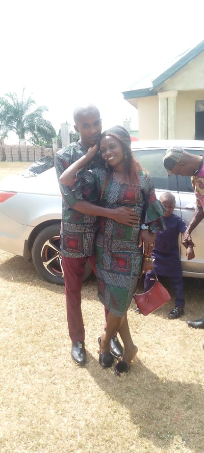 A smiling couple in matching patterned outfits stands outdoors. The man wears trousers, while the woman wears a dress and carries a purse. They are embracing in front of a silver car. In the background, there's a young child in a blue outfit and an older person wearing a patterned shirt. Stacks of bricks and a building can also be seen. The ground is covered in dry grass.