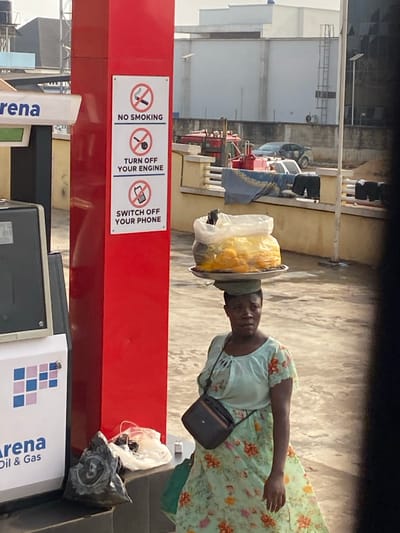 The image captures an outdoor scene in Issele Uku, Nigeria, under bright daylight. A dark-skinned woman is positioned in the midground, slightly to the right of the frame's center, facing partially towards the right. She wears a light green dress with a floral pattern of orange, green, and pink, and has a small black rectangular purse with a strap slung across her body. She balances a round, metallic tray on her head, which holds a large clear plastic bag filled with numerous yellow or orange spherical fruits, and a dark, unidentifiable object. Her head is covered by a green cloth.
To the left of the woman, a red vertical pillar is prominent. Affixed to this pillar are three white rectangular signs, vertically aligned, each featuring a safety directive with an accompanying icon: "NO SMOKING" with a crossed-out cigarette, "TURN OFF YOUR ENGINE" with a crossed-out car key, and "SWITCH OFF YOUR PHONE" with a crossed-out mobile phone. Partially visible to the immediate left of the red pillar is a white and grey gas pump labeled "Arena Oil & Gas." At the base of the pillar, on a low concrete block, a black plastic bag and a clear plastic bag rest.
In the background, a low, light-colored concrete wall with railings is visible. Draped over this wall are articles of clothing, including a blue blanket and dark trousers. Beyond the wall, several vehicles are present: a red tractor, a silver sedan, and another dark-colored car. Further in the distance, light-colored buildings with flat roofs stand against a clear sky. The ground throughout the visible area appears to be paved.