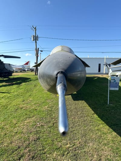 The nose of a dark-colored aircraft, possibly a helicopter, dominates the foreground. A silver probe extends forward from the nose cone. Lush green grass covers the ground, with a building, power lines, and additional aircraft visible in the blurred background. The sky is a clear blue, and the overall composition suggests an outdoor aviation museum or display.