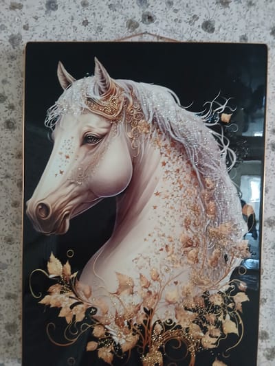 This media file is a close-up photograph of a framed artwork, likely a painting or a mixed-media piece, depicting a stylized, ethereal horse. The horse is rendered in shades of white and light brown, with a flowing mane adorned with what appear to be tiny beads and golden leaves or floral elements. The horse's head is turned to the left, with its eye gazing forward. There are star-shaped embellishments on its muzzle and forehead. The background of the artwork is a stark black, which makes the light-colored horse and golden accents stand out prominently. The artwork is framed by a thin, gold-colored border. The photograph is taken from a slight angle, showing the texture of the wallpaper behind the artwork, which has a delicate floral pattern in shades of gray and white. The lighting suggests it is indoors, possibly in a home or gallery setting. There is no visible text in the image. The overall impression of the artwork is one of fantasy, grace, and a touch of magic.
