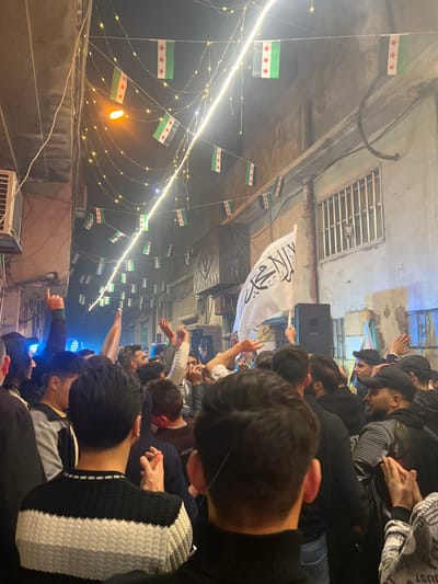 A group of people gathered under an awning or ceiling decorated with Syrian flags and string lights. Some hold up a white flag with Arabic text. They appear to be celebrating or protesting. Some have their hands raised in the air or are making gestures. A speaker is visible, suggesting music or amplified sound. The scene seems to be in an urban area.