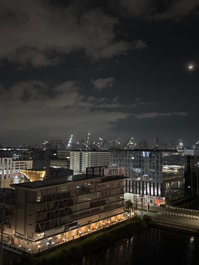 The image depicts an urban night scene in Pasay City, Philippines. In the foreground, a dark body of water, likely a river or canal, is visible. A bridge spans the water, connecting to a road. To the left of the water and bridge is a multi-story building with glass windows, featuring prominent lighting at its base and illuminated floors. A sign with the word "JETTI" is affixed to the side of this building. Behind this structure, a cityscape stretches into the distance, characterized by numerous tall buildings and skyscrapers. Construction cranes, illuminated with green lights, are visible among the buildings, suggesting ongoing development. The sky is dark, with scattered clouds, and a sliver of a crescent moon is present in the upper right quadrant of the image. The overall impression is that of a bustling, modern urban environment at night.