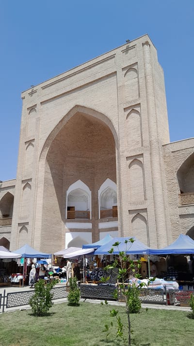 A low-angle, full shot captures a bustling outdoor market in Denov, Uzbekistan, set against the backdrop of a large, pale brick architectural structure with multiple archways and balconies. The sky is a clear, bright blue. In the foreground, a neatly mowed green lawn is dotted with small, leafy green trees. A black metal fence with an intricate geometric pattern separates the lawn from the market stalls.
Several market stalls, covered by colorful awnings (predominantly blue and white), display a variety of goods. Items visible include clothing, textiles, and what appear to be souvenirs or handicrafts. People are seen browsing and tending to the stalls. One stall features stacks of patterned fabrics and what look like pillows. Two individuals stand near a stall, with one appearing to be a vendor. Another person is visible further back, closer to the large brick building.
The monumental brick building dominates the upper portion of the image. Its facade features a massive central archway, with smaller arched niches and balconies visible on either side. The brickwork is light in color, and decorative elements, such as vertical ribbed patterns and recessed rectangular shapes, are integrated into the design. The architectural style suggests a historical or cultural significance to the location. The overall scene portrays a vibrant commercial activity taking place in front of a historically impressive building under a sunny, clear sky.