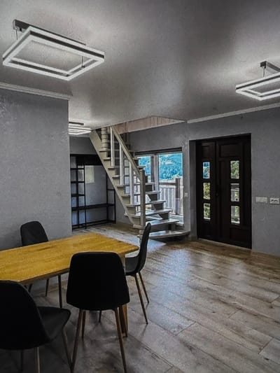 The image shows the interior of a modern home, likely a living or dining area, with a view of a mountainous landscape through a large window and a staircase leading up. The room has grey walls and light-colored wooden flooring. In the foreground, a rectangular wooden dining table is set with four black chairs. A large, dark wooden double door is visible on the right side of the room, and to the left of it, a set of light-colored stairs ascends. Above the dining area, two rectangular pendant light fixtures hang from the ceiling. The view through the window reveals a lush green mountainside with trees. The scene does not provide any specific indicators of location within the city of Dubno, Ukraine.