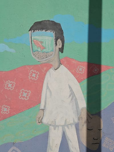 The image is of a mural, likely located in Osorno, Chile, showing a figure lying horizontally against a painted backdrop of blue, green, and lavender. The figure is painted in shades of white and grey, wearing what appears to be a long-sleeved garment. The head of the figure is depicted as an open television screen displaying a red fish swimming in a blue, watery environment. The rest of the figure appears to be a regular human head. The painted backdrop behind the figure includes geometric patterns and abstract color fields. The mural's surface has cracks and imperfections, typical of an outdoor wall. A dark shadow passes across the image, obscuring the upper section of the painting.