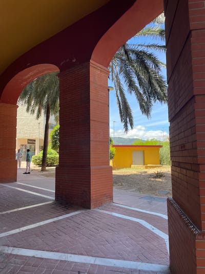 This image shows a view through an archway onto a courtyard with palm trees and a yellow building. Two people are visible in the distance, standing on a brick pathway. The sky is blue with some clouds. The overall scene appears to be outdoors, likely in a warm climate given the presence of palm trees. The architecture suggests a possibly Mediterranean or Spanish setting, which aligns with the location context of Algeciras, Spain. The time of day appears to be daytime, with bright sunlight casting shadows. There is no visible text in the image.