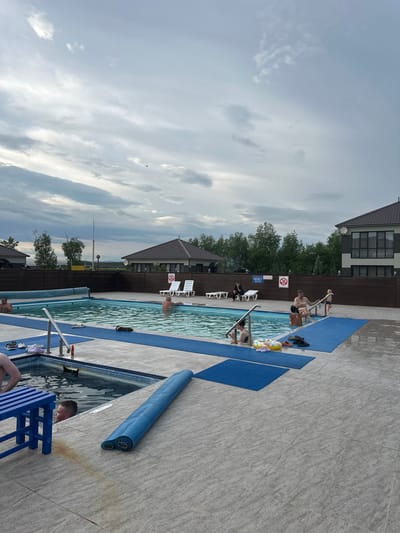 The image depicts an outdoor swimming pool area, likely in a resort or private residence setting. The pool is rectangular and filled with clear water. Several people are visible in and around the pool. In the main pool, two men are partially submerged, with one appearing to be standing and the other leaning against the side. A child is entering the pool from the steps, accompanied by an adult woman. Another woman is seated on the edge of the pool, interacting with a young child. Two lounge chairs are placed on the tiled area surrounding the pool, with two people seated on them. A blue bench is positioned near a smaller, square pool which also contains water and a child.
The pool area is paved with light-colored tiles, with blue mats laid out in sections. Objects such as flip-flops, a rolled-up blue mat, and a yellow floatation device are scattered on the ground. In the background, there are modern houses with dark roofs and visible windows, surrounded by a wooden fence and trees. The sky is overcast with dramatic cloud formations. The scene suggests a relaxed, recreational atmosphere, possibly during a summer day, given the presence of swimwear and swimming activities.