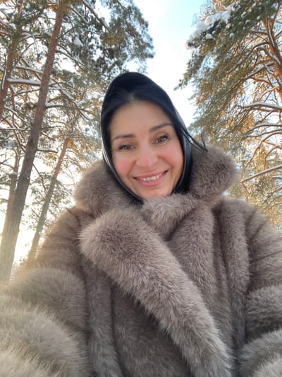 The image presents a close-up selfie of an adult female in a snowy forest environment in Syasstroy, Russia. The subject, viewed from the upper chest upwards, has dark hair partially covered by a black head covering. She is wearing a thick, light brown/grey fur coat featuring a large collar. Her facial expression displays a broad smile, with her eyes directed toward the camera. The background consists of numerous tall evergreen trees, their branches visibly laden with snow, under a bright, clear sky. Sunlight emanates from the left side of the frame, illuminating portions of the snow-covered foliage.