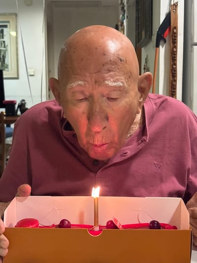 A forensic examination of the image reveals an elderly, bald male, likely in a private residence in Bnei Dror, Israel, engaged in the act of blowing out a single candle. The subject, seen from the chest up, wears a maroon collared polo shirt with a faint embroidered emblem on the left chest. His eyes are closed, and his lips are pursed, indicating exhalation towards the candle. He holds a brown and white rectangular dessert box containing a single lit, gold-colored candle. Inside the box, red decorative elements resemble ribbons, accompanied by three dark red spherical items, likely cherries, and a small white tag. The candle's flame casts an orange luminescence on the lower part of his face. The background displays domestic elements: a framed picture and white streamers on the left, and a vertical carved wooden ornament alongside a dark utility handle on the right, suggesting an interior household setting.