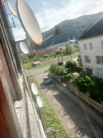 The image shows a collection of satellite dishes mounted on the side of a building, viewed from a high angle. The satellite dishes are of various sizes, with one large, prominent dish in the foreground angled towards the sky. Several smaller dishes are visible further back, attached to the building's exterior. The scene is set outdoors, likely on a balcony or window ledge overlooking a street. Below the building, a road can be seen with a parked car and some greenery. The background features a lush, tree-covered hillside under a bright, cloudy sky, suggesting it's daytime. The building itself appears to be multi-story, with visible windows and a white facade. There is no discernible activity or event taking place beyond the static presence of the satellite dishes and the surrounding environment. The image is taken from a perspective that emphasizes the satellite equipment against the natural landscape. The orientation of the photo is sideways.