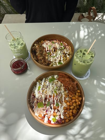 The image depicts an outdoor dining scene at Café Sinónimo, located at Calle Gregorio Dávila 71, Americana, 44160 Guadalajara, Jalisco, Mexico. On a light-colored table, two large wooden bowls containing chilaquiles are centrally positioned. Each bowl features tortilla pieces covered in red and green sauces, drizzled with white crema, and garnished with crumbled cheese, chopped red onion, and cilantro sprigs. The upper bowl contains shredded brown meat on its left side. The lower bowl includes shredded reddish meat on its upper right side and roasted chickpeas on its lower right side. Flanking the food are two clear glasses filled with iced green beverages, likely matcha lattes, each with a wooden straw and resting on white coasters bearing the "CAFÉ SINÓNIMO" logo. A small clear jar of red condiment is present to the left of the upper food bowl. In the upper background, a person wearing a black long-sleeved shirt is partially visible, alongside a light-colored woven straw bag to their right. Dappled sunlight casts shadows across the table, and out-of-focus green foliage is visible in the background.