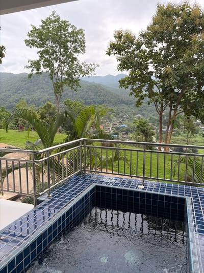 A view of a small tiled pool overlooking a lush green valley and a village in Ban Kao Dua, Thailand. The pool has dark blue tiles and is filled with water. A metal railing surrounds the pool. In the background, there are rolling hills covered with trees and a few houses scattered throughout the landscape. Several large trees with green foliage are visible in the foreground and midground. The sky is overcast.
