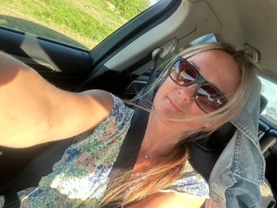 A woman is shown from the chest up, inside a vehicle. She is wearing sunglasses and a floral print dress. Her hair is blonde and tied up. A denim jacket is draped over the headrest of the car seat behind her. Sunlight is illuminating the scene, casting shadows on the interior of the car and the woman's arm. The background outside the car window shows greenery and a clear sky. The specific location in Novotolucheyevo, Russia, is not discernible from the image.