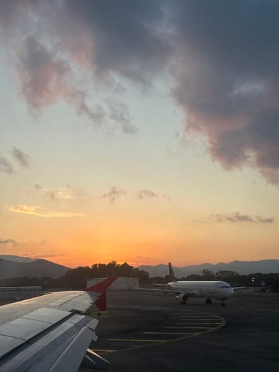 The image depicts an airport scene at sunset, observed from the window of an aircraft, presumably at Dalaman Airport in Dalaman, Turkey. The sky is dominated by a vibrant gradient of colors, transitioning from deep orange, red, and yellow at the horizon, where the sun is setting, to lighter blue and grey-pink in the upper portions. Scattered clouds, some tinged with pink and purple, are present across the sky.

In the lower left quadrant, the silver-grey wing of the observer's airplane is prominently featured, displaying its flaps and a red wingtip fence. The wing is sharply detailed, with visible panel lines and structural elements.

In the midground, on the tarmac to the right, another white narrow-body passenger aircraft, likely an Airbus A320 series, is stationary. It is oriented with its nose slightly turned towards the viewer, revealing its cockpit windows and two underwing engines. Yellow painted lines, indicative of taxiways or parking positions, are visible on the dark grey asphalt tarmac around this aircraft.

The background features a range of dark, silhouetted mountains stretching across the horizon. Below the mountains, obscured by the low light, are dark trees and various airport infrastructure elements, including what appear to be industrial tanks and buildings. No individuals are visible in the scene. The overall lighting suggests the transition from late afternoon to evening.