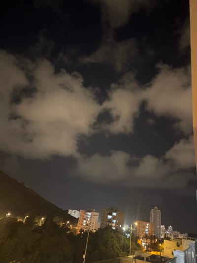 This is a nighttime photograph of a cityscape in Tirat Karmel, Israel, taken from a higher vantage point, likely a balcony or elevated structure. The sky is dark with scattered clouds illuminated by city lights. In the foreground, to the left, a steep, tree-covered hillside slopes downwards, with several streetlights visible along its path. Beyond the hillside, a cluster of multi-story residential buildings with illuminated windows are situated. The buildings vary in height and architectural style, with one taller, slender building standing out among them. The overall impression is a residential area at night, characterized by the contrast between the dark natural landscape and the lit-up urban structures.
