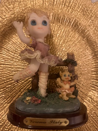A close-up shot shows a figurine of a young ballerina in a pink tutu and ballet slippers standing on a grassy base. A small, anthropomorphic cat character, also in a ballet costume, stands to the right of the ballerina. The figurine is placed on a textured gold background, possibly a decorative plate. A wooden base with a plaque that reads "Verona Italy" is visible at the bottom of the figurine. The scene is lit with warm, artificial light. There is no indication of the city of Edem, Russia, in the image.