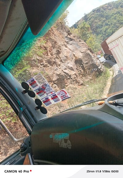 From the driver's seat, a winding mountain road unfolds. A large rocky cliff dominates the view, partially obscured by the windshield and its affixed stickers. Another vehicle, possibly a truck, is visible on the narrow road ahead. The dashboard, with signs of wear, fills the lower portion of the frame. The sky appears hazy, suggesting a sunny day.
