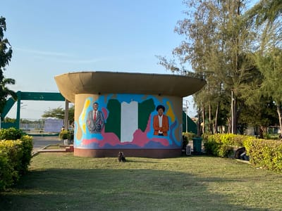 The image shows a circular, dome-like structure with a mural depicting the map of Nigeria and portraits of two individuals in Tawpo, Nigeria. The mural is painted on the side of the structure. The Nigerian flag forms the center of the map. To the left of the map is a man in a gray suit with a red tie, and to the right is a woman in a brown suit. The structure is on a grassy area, with bushes and trees in the background. A person is sitting near the bushes to the right of the structure. There is a green trash can next to the structure. In the far background is a green and white gate structure. The sky is blue and clear. A dark brown stump is present near the base of the structure.