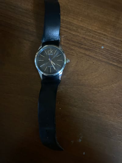 An overhead, close-up shot features a black leather-strapped Calvin Klein watch resting on a wooden surface. The watch face is dark gray with silver hour markers and hands. The "ck" logo and "Calvin Klein" text are visible at the top of the face, with "Swiss Made" below the center. The watch is slightly angled on the wooden grain, which has visible lines and some specks of lighter material.
