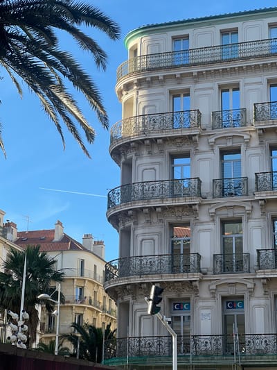 The image depicts a view of a curved building facade with ornate balconies and windows, typical of Haussmannian architecture. The building is light-colored, with decorative iron railings on the balconies. Several windows are visible, reflecting the clear blue sky. In the foreground, a portion of a palm tree frond extends into the frame from the left. In the background, another building with a red tiled roof and chimneys can be seen. A contrail from an airplane is visible in the sky. A traffic light and a surveillance camera are also present in the foreground. The location is identified as Toulon, France.