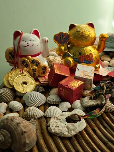 The image displays a collection of decorative items, primarily two "maneki-neko" (beckoning cat) figurines and several small boxes, all arranged on a textured surface that appears to be made of woven reeds or wood. The background is a plain, light green wall.
In the foreground and scattered around the figurines are numerous seashells of various shapes and sizes, as well as some pieces of coral or weathered shells.
The left figurine is a white maneki-neko with red accents, holding several gold-colored coins on strings. The right figurine is a larger, golden maneki-neko, also holding decorative elements. Between the figurines are several small, red boxes with gold imprints, and one white box with gold patterns. A colorful, patterned ribbon or fabric is draped across some of the shells on the right side of the frame.
There is no indication of the city of Sihanoukville, Cambodia, or any specific geographical location within the image itself. The scene is a still life composition of decorative objects.