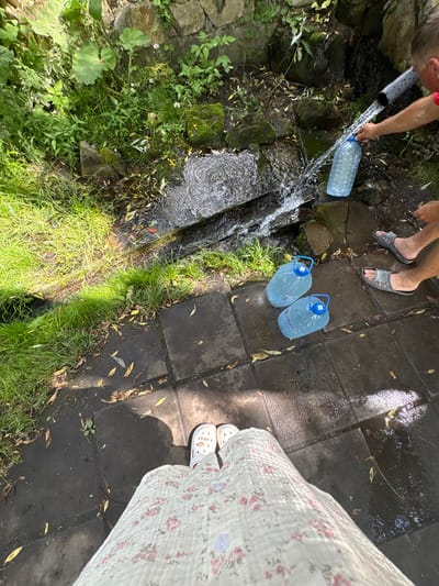 The image depicts an outdoor scene near a water source, likely a spring, in Новоподклетное, Russia. In the upper right, a person's arm and shoulder are visible, holding a grey pipe from which water is flowing into a clear plastic bottle. In the foreground, three large clear plastic water bottles with blue handles are placed on a dark tiled surface. Two of these bottles are full, and one is partially filled. Nearby, two pairs of feet wearing grey sandals are visible on the tiles. In the bottom foreground, the legs of a person wearing a white floral-patterned dress and white clogs are shown, also standing on the tiled area. The environment includes lush green grass, foliage, rocks, and a narrow channel of flowing water. The ground is wet, indicating the presence of water.