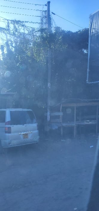 A hazy outdoor shot reveals a street scene. A white van is parked on the right side of the road. Behind the van, what appears to be a metal scaffolding structure stands alongside a tree. An electrical pole with multiple wires attached is visible on the left. The sky is light blue and mostly clear.