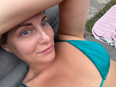 The image shows a close-up, overhead shot of a woman lying on a grey lounge chair, with her arm raised above her head. She is wearing a teal bikini. To the right of the chair, a pink and white striped towel is visible on green grass. The scene appears to be outdoors, likely in a backyard or garden area. No specific landmarks or indicators of the city of Rueil-Malmaison, France are present in the image.